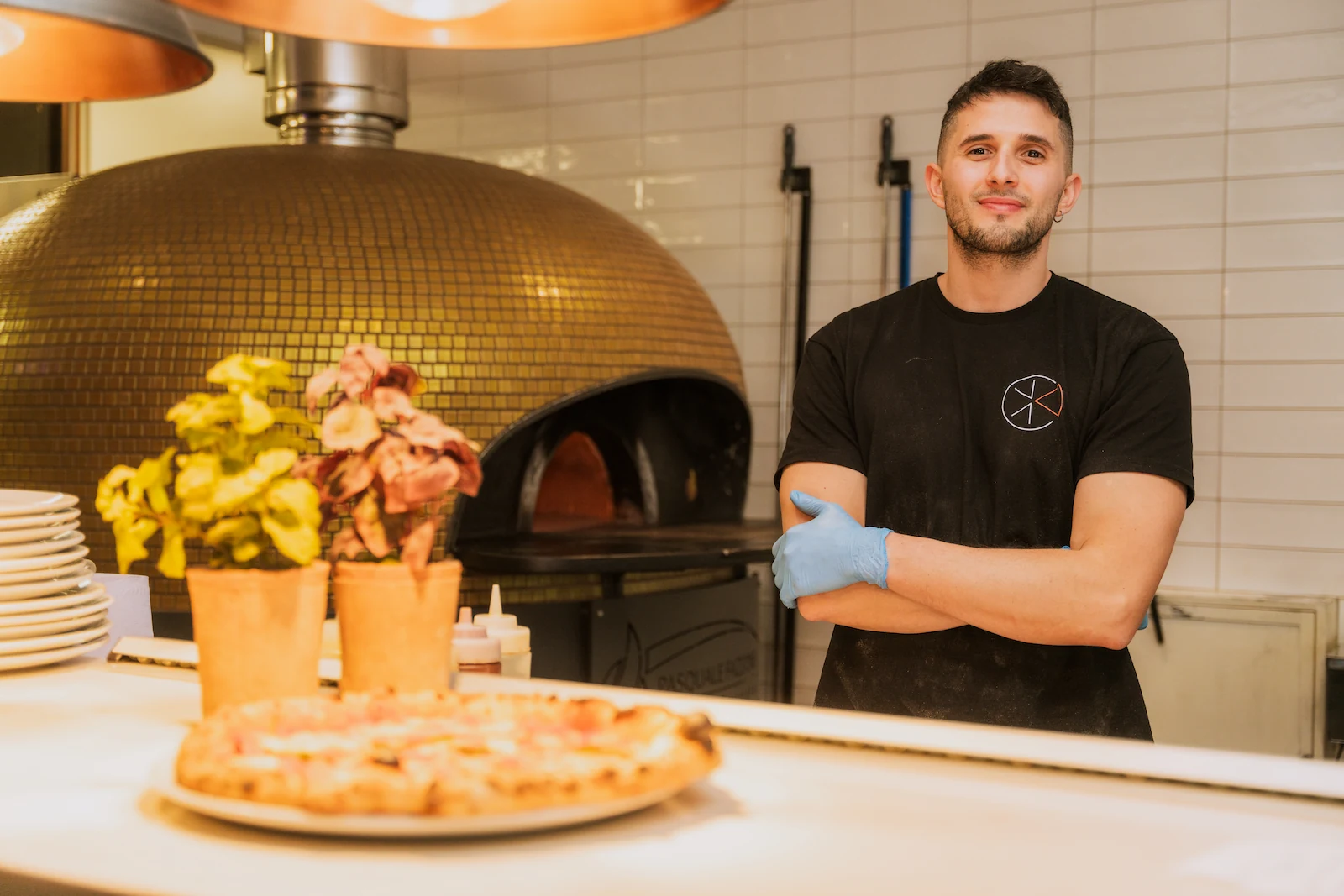 A chef is smiling with a cooked pizza in the foreground.