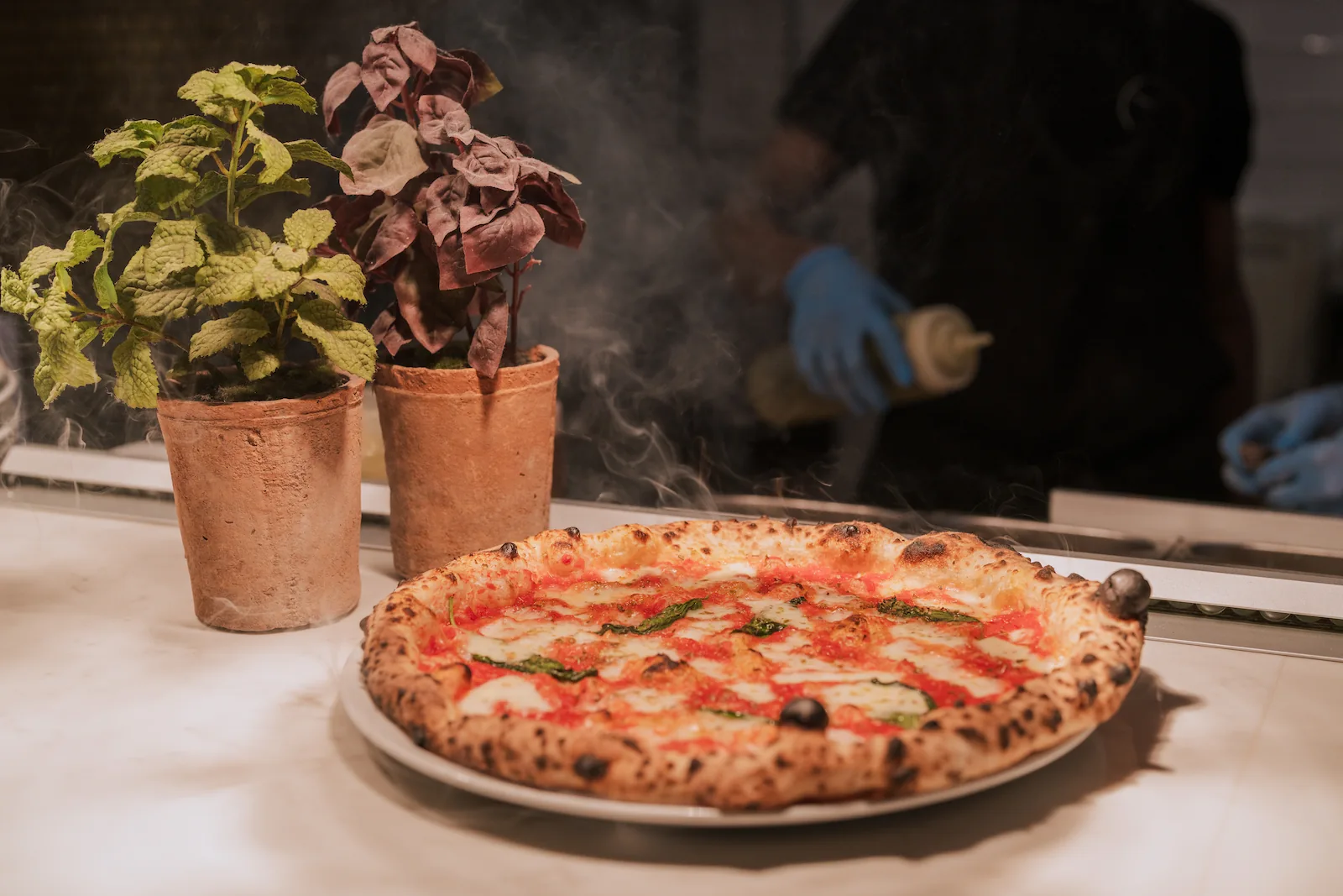 A cooked pizza is steaming with a chef in the background and two plant pots.